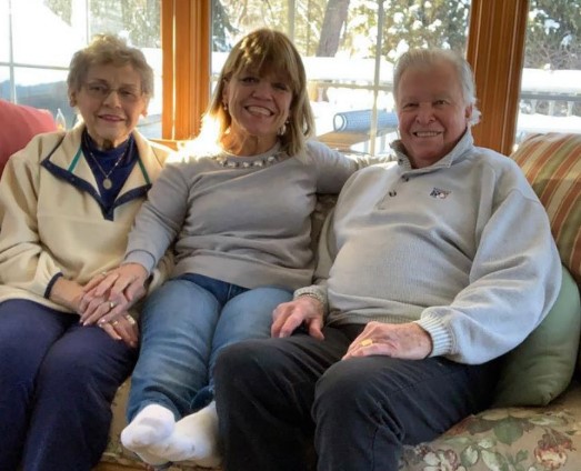 Amy with her parents: Mother on the left, father on the right, while Amy is sitting between them on a sofa on their living area.