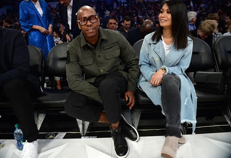 Dave and his wife sitting during a game at the NBA All-Star Game 2018 at Staples Center on February 18, 2018 in Los Angeles, California. In a dark green shirt is Dave with one of his arms under his lift thigh and Elaine is in a blue trenchcoat smiling with her hands cusped together over crossed legs.