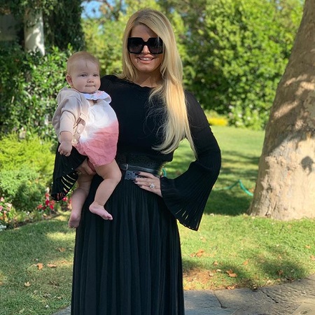 Thin Jessica Simpson in black dress with her left hand on her hip while holding her daughter in her right hand.