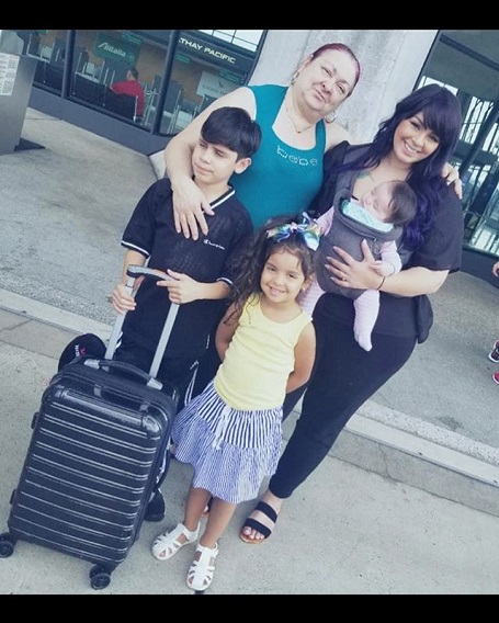 Tiffany Franco holding her daughter in a holding crib while her mother put her arm on her. Two kids stand in front of them, her son holding a bag's handle.