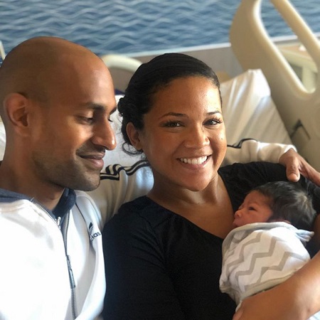 Tony looking at the baby in Laura's arm while she is smiling at the camera. They're sitting on a bed.