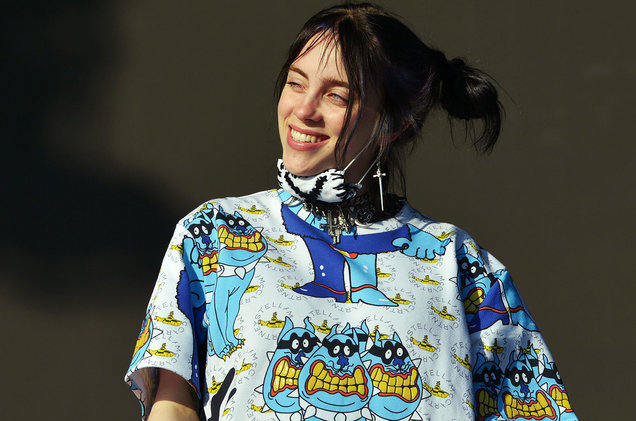 billie on stage in the coachlella event with space buns, mask and a cartoon printed oversized shirt smiling at her fans 