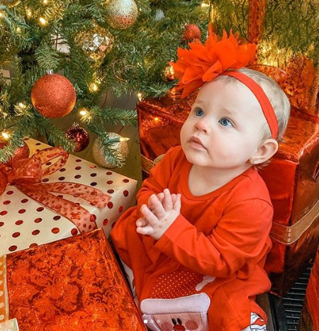 Baby Mila under a christmas tree dressed up in a red bodysuit.