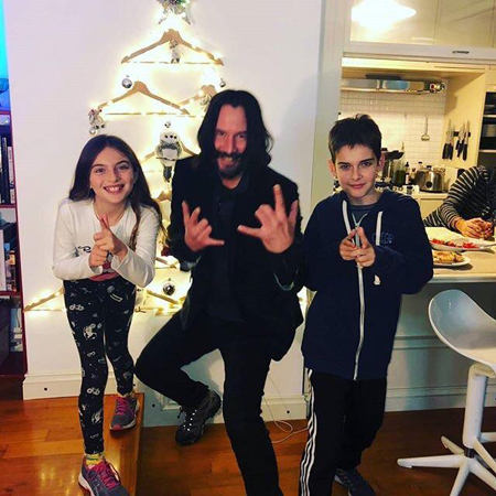 Keanu, with two kids on his left and right smile and pose for the camera.