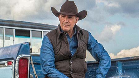 Kevin Costner sits behind a pick up truck with a cowboy hat.