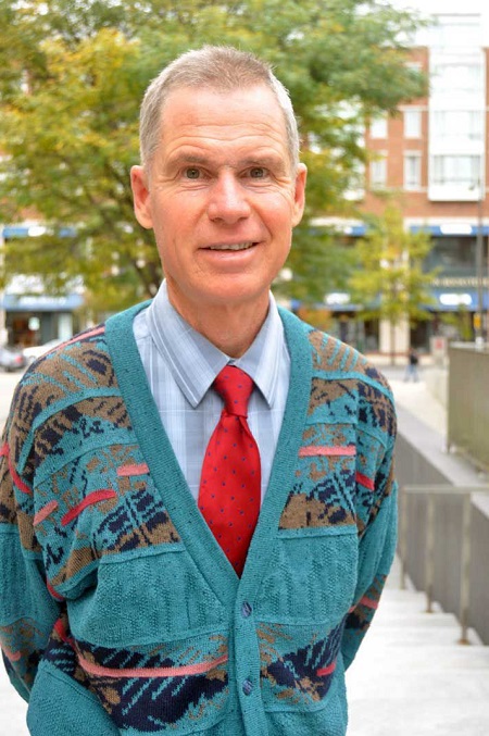 John Rodden wearing a sweater outside a shirt and a tie, posing with his hands back.